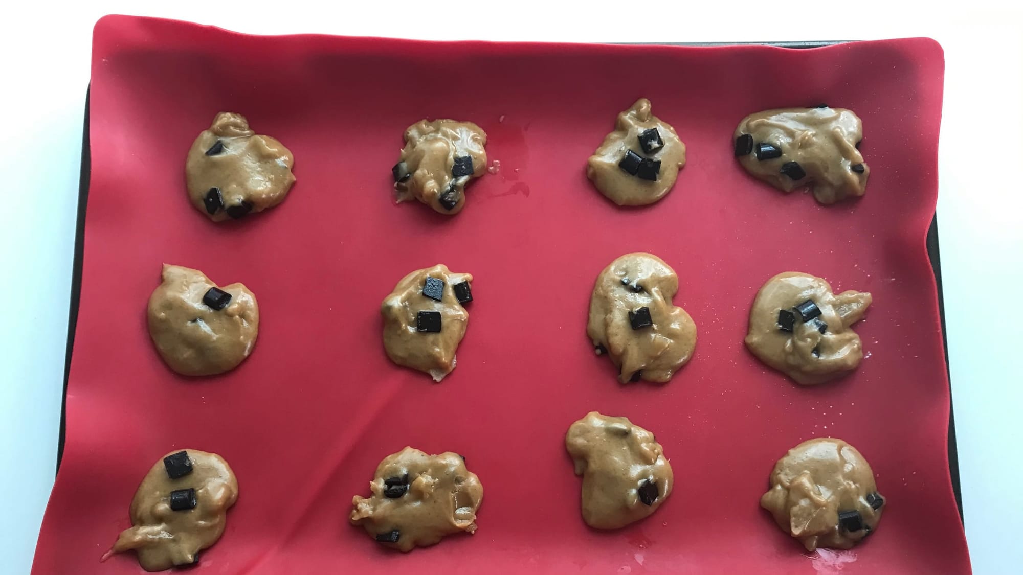 12 glistening cookies before baking, showing the wet dough and spacing suggestion, laid out on a red silicone baking tray