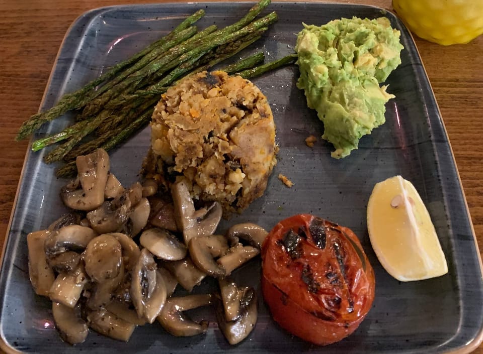 A square plate with separate portions of grilled mushrooms, tomato, vegan bubble & squeak, guacamole, and asparagus.